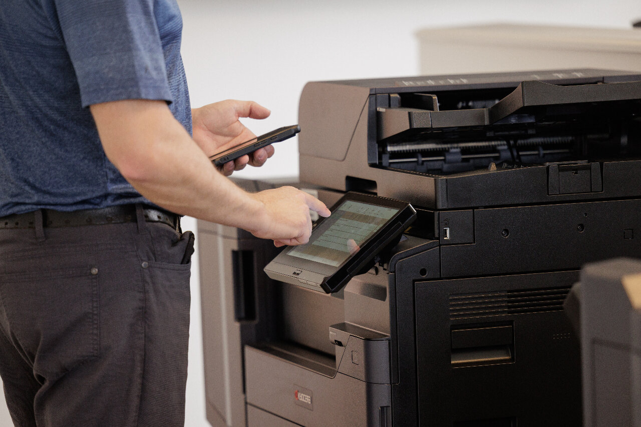 Service Technician standing at Kyocera Multifunction Printer with mobile device in hand sending a document to the device via mobile print